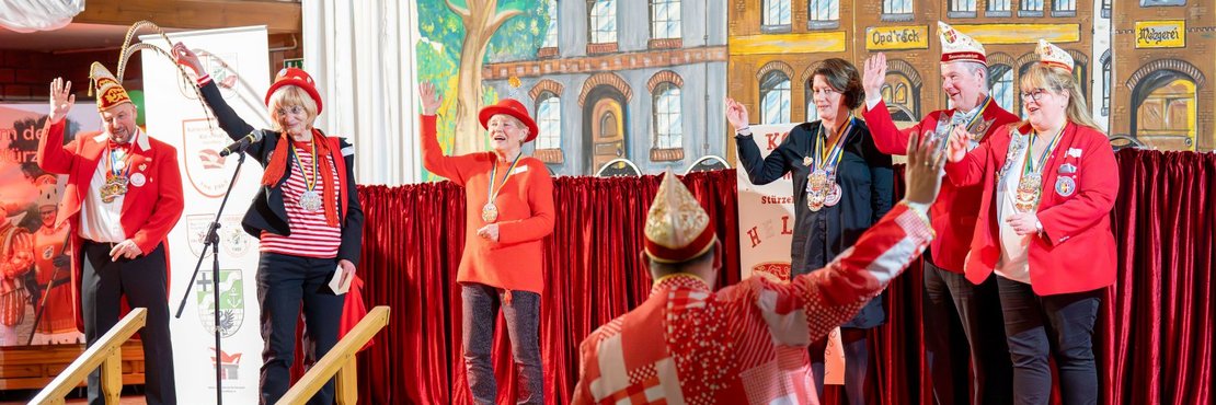 Auf der Bühne stehen sechs Personen, die alle lächeln und mit einer Hand ins Publikum winken. Die Personen sind festlich gekleidet, viele tragen rote Jacken, Hüte und Karnevalsorden. Hinter ihnen befindet sich ein Bühnenbild, das eine stilisierte Stadtansicht mit gemalten Häusern und einem Baum zeigt. Der Hintergrund ist mit einem schweren roten Vorhang verziert.