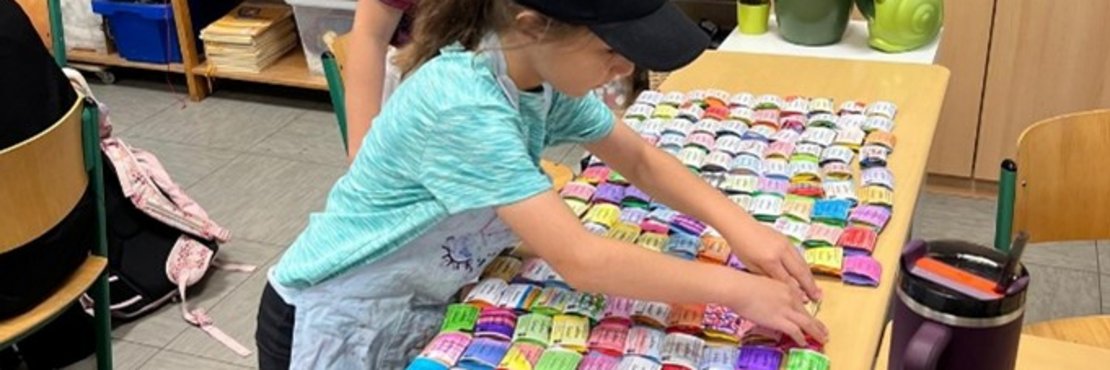 Zwei Mädchen arbeiten an Schreibtischen in einem Klassenzimmer und sortieren farbenfrohe, beschriftete Quadrate, die auf den Tischen ausgebreitet sind.