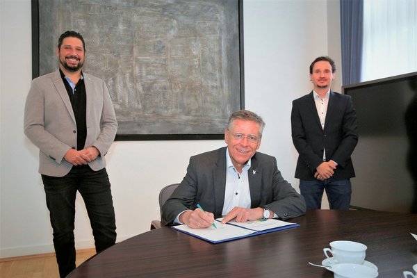 Landrat Hans-Jürgen Petrauschke sitzt am Tisch und unterschreibt Dokument. Dahinter stehen Thiago de Carvalho Zakrzewski und Fabian Tenk.