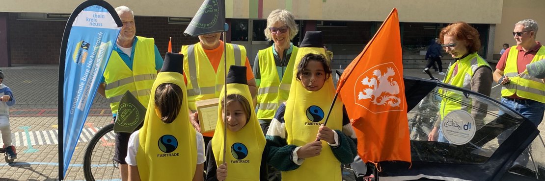 Drei Kinder in gelben Bananen-Kostümen mit Fairtrade-Logo stehen im Mittelpunkt vor einem Schulgebäude. Eines hält eine orangefarbene Flagge mit weißem Löwenwappen. Dahinter stehen mehrere Erwachsene in gelben Warnwesten. Links eine blaue Fahne der 'Grundschule Burg Hackenbroich – wir sind Fairtrade School' vom Rhein-Kreis Neuss. Rechts ein schwarzes Lastenrad mit Dormagen-Logo.