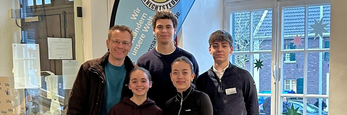 Das Bild zeigt eine Gruppe von fünf Personen, wahrscheinlich Schüler und ein Lehrer, die hinter einem Stand stehen. Der Stand ist mit einer Tischdecke bedeckt, die teilweise mit einer grünen Stoffbahn mit der Aufschrift "Fairtrade-School" verziert ist. Auf dem Stand befinden sich verschiedene fair gehandelte Produkte, darunter Schokolade und Nüsse. Ein Schild wirbt mit "Zeit für faire Schokolade!". Im Hintergrund ist ein Banner der "Fair Trade Company Knechtsteden" zu sehen. Die Szene deutet auf eine Verkaufsaktion oder einen Informationsstand zum Thema Fair Trade hin.