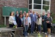 Das Bild zeigt eine Gruppe von etwa 15 Personen, die vor einer Backsteinmauer stehen.  Im Hintergrund steht eine grüne Tafel, auf der mit Kreide „Hallo“ und „Ne…“ (vermutlich unvollständig) geschrieben steht.  Die Personen wirken wie eine Gruppe von Erwachsenen, möglicherweise ein Arbeitsteam oder eine politische Gruppe, die sich für ein Foto versammelt haben.  Manche halten Dokumente oder Getränke.  Der Ort scheint ein Außenbereich zu sein, mit Gartenelementen wie Pflanzen und Tischen mit Stühlen im Vordergrund. Die Atmosphäre ist freundlich und ungezwungen.
