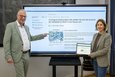 Zwei Personen, ein Mann und eine Frau, stehen vor einem großen, auf einem Standfuß stehenden Touchscreen. Der Mann, links im Bild, trägt einen hellgrünen Anzug, ein hellblaues Hemd und eine Brille. Er hat kurze, graue Haare und zeigt mit dem Finger auf den Bildschirm. Die Frau, rechts im Bild, trägt einen grauen Blazer und eine blaue Hose. Sie hält ein Tablet in der Hand, auf dem die gleiche Umfrage wie auf dem Bildschirm zu sehen ist. Sie hat lange, braune Haare und ein freundliches Lächeln im Gesicht. Der Bildschirm zeigt eine Webseite mit der Überschrift „Umfrage & Gewinnspiel: Wie stellen Sie sich die Zukunft der Mobilität im Rhein-Kreis Neuss vor?".  Es gibt verschiedene Schaltflächen, darunter „Informationen", „Übersicht", „Umfrage", „Rhein-Kreis Neuss" und „Verkehr und Mobilität". Unter der Überschrift ist ein Bild einer Stadtkarte mit verschiedenen Verkehrsmitteln zu sehen. Der Text auf dem Bildschirm beschreibt eine Bürgerbeteiligung zur Entwicklung eines integrierten Mobilitätskonzeptes bis 2045 und ruft zur Teilnahme an einer Online-Umfrage auf, mit der Möglichkeit, einen Einkaufsgutschein zu gewinnen.