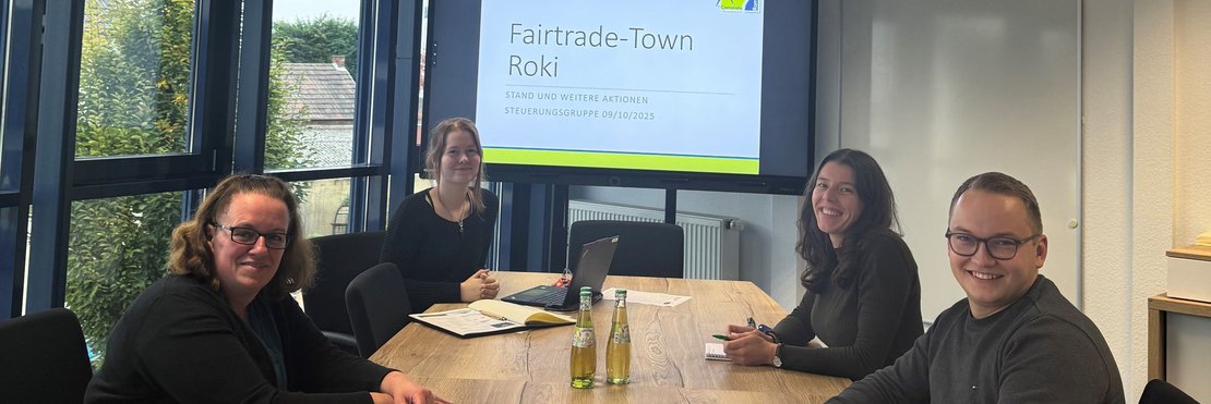 Drei weibliche und eine männliche Person sitzen an einem langen Tisch. Im Hintergrund ist eine Präsentation mit dem Titel "Fairtrade Town Roki" zu sehen