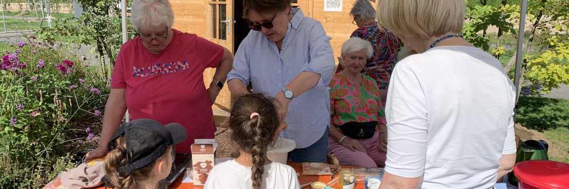 Eine Gruppe von Menschen steht um einen Tisch im Freien. Sie bereiten Lebensmittel zu die auf dem Tisch liegen.