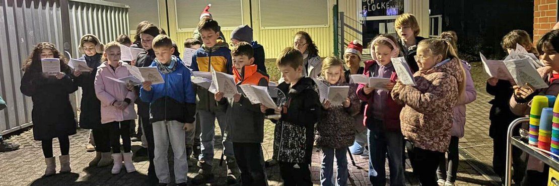 Das Bild zeigt eine Gruppe Kinder, die im Freien stehen und Weihnachtslieder singen. Sie halten Notenblätter in den Händen und sind in winterliche Kleidung gehüllt. Im Hintergrund sind Container und Bäume zu sehen.