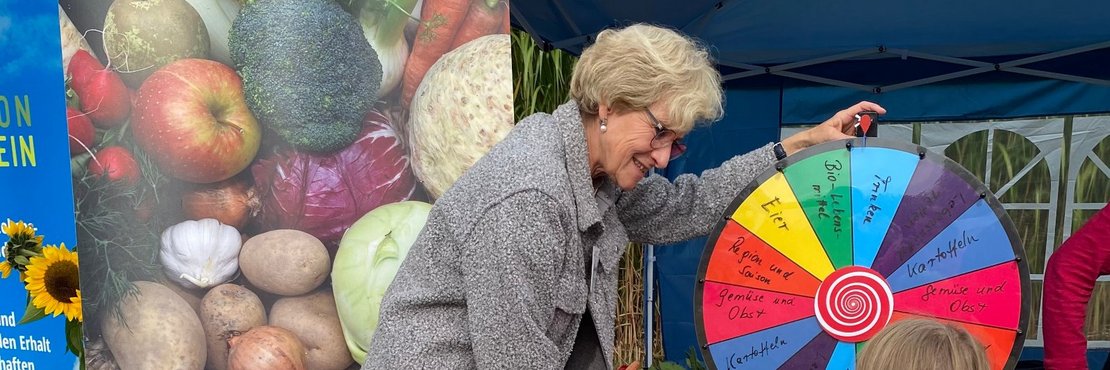 Das Bild zeigt eine ältere Frau mit kurzen, blonden Haaren, die sich über ein buntes Glücksrad beugt. Das Rad ist in verschiedene farbige Segmente unterteilt, jedes mit einem regionalen Lebensmittel beschriftet, wie z.B. Eier, Gemüse und Obst, Kartoffeln, Bio-Lebensmittel und Getränke. Die Frau trägt eine graue Jacke und scheint fröhlich und engagiert zu sein. Neben ihr steht ein kleineres Kind in einem rosa Oberteil, das gespannt auf das Ergebnis des Drehens schaut. Vor dem Glücksrad steht ein kleiner Korb mit einigen Früchten und Gemüse. Im Hintergrund ist ein großer Banner zu sehen, der mit einer bunten Darstellung von regionalem Obst und Gemüse dekoriert ist. Der Banner selbst wirbt mit dem Slogan "Regional essen! Mach mit!".