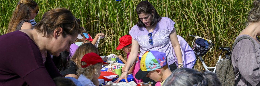 Mehrere Kinder und Erwachsene sitzen an langen Tischen und gestalten mit bunten Filzstiften Stoffbeutel. Im Hintergrund stehen zwei blaue Luftballons vor hohem grünen Schilf.