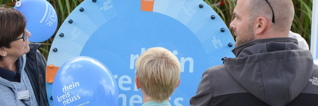 Das Bild zeigt eine Frau mit kurzen, dunklen Haaren und einer dunkelblauen Jacke, die sich links im Bild befindet. Sie trägt eine Brille und scheint mit Interesse auf das Glücksrad zu schauen. Neben ihr schwebt ein blauer Luftballon mit dem Aufdruck „Rhein Kreis Neuss“ und der dazugehörigen Webseite. Im Mittelpunkt des Bildes steht ein Junge mit blonden Haaren, der auf das Glücksrad schaut und ein hellblaues T-Shirt trägt. Rechts im Bild steht ein Mann mit kurz geschnittenen Haaren, einer dunklen Jacke mit aufgestelltem Kragen und Sonnenbrille auf dem Kopf. Hinter den Personen ist hohes Gras zu sehen.