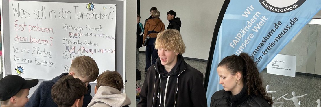 Im Vordergrund des Bildes stehen mehrere Schüler, die sich um eine große, weiße Tafel versammelt haben. Die Tafel trägt die Überschrift "Was soll in den Fair-o-maten?" und darunter Anweisungen wie "Erst probieren, dann bewerten!" und "Verteile 2 Punkte für deine Favoriten". Auf der Tafel sind verschiedene Produkte aufgelistet, darunter "Mango-Stripes", "Schoko-Cookies" und "Nüsse", jeweils mit vielen waagerechten Strichen, die Stimmen darstellen. Die Schüler sind überwiegend in Freizeitkleidung gekleidet, darunter Jacken und Hoodies in verschiedenen Farben wie Blau, Grau, Braun und Schwarz.