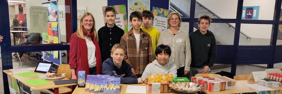 Das Bild zeigt eine Gruppe von Schülern und zwei Lehrerinnen an einem Stand innerhalb eines Schulgebäudes. Die Schüler stehen hinter dem Tisch, auf dem verschiedene Fairtrade-Artikel ausgestellt sind, darunter Lebensmittel wie Kekse, Getränke und Süßigkeiten. Ein auf dem Tisch ausgebreitetes Poster zeigt eine Weltkarte mit Informationen zu Ernährung und Fairtrade. Auf einem anderen Plakat steht "Nikolaus Tombola" geschrieben.