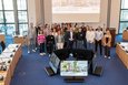Das Bild zeigt eine Gruppenaufnahme von etwa 30 Personen, die im Kreissitzungssaal in Grevenbroich stehen. Im Hintergrund ist eine Leinwand mit einer Präsentation zu sehen, deren Titel „Grundlagenwissen - „Nachhaltigkeit in der Stadtplanung / Nachhaltiges Bauen" lautet. Die Personen scheinen an einer Veranstaltung oder einem Seminar zum Thema nachhaltiges Bauen teilzunehmen. Die Stimmung wirkt freundlich und konzentriert. Die Personen sind verschieden alt und tragen unterschiedliche Kleidung. Vorne stehen zwei Bildschirme mit weiteren Informationen zum Thema.