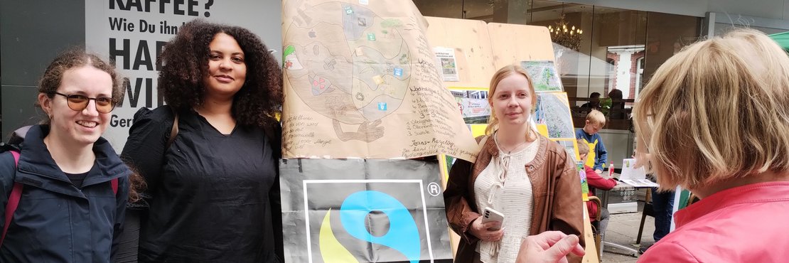 Drei Frauen stehen neben einem Plakat von "Fairtrade" an einem Marktstand.