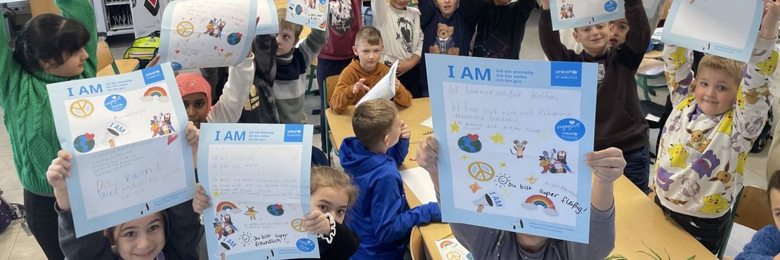 Das Bild zeigt eine Gruppe von Kindern in einem Klassenzimmer. Sie halten alle ein Blatt Papier hoch, auf dem "I AM" steht und das mit Zeichnungen und Texten versehen ist. Der Hintergrund des Bildes ist ein Klassenzimmer mit Regalen, Schreibtischen und Stühlen. Auf den Regalen befinden sich verschiedene Gegenstände, darunter Bücher, Ordner und Spielzeug.