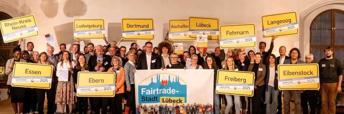 Eine Gruppe an Menschen stehen auf einer Bühne und halten Schilder in der Hand. Auf den Schildern stehen die Städtenamen der Mitglieder.