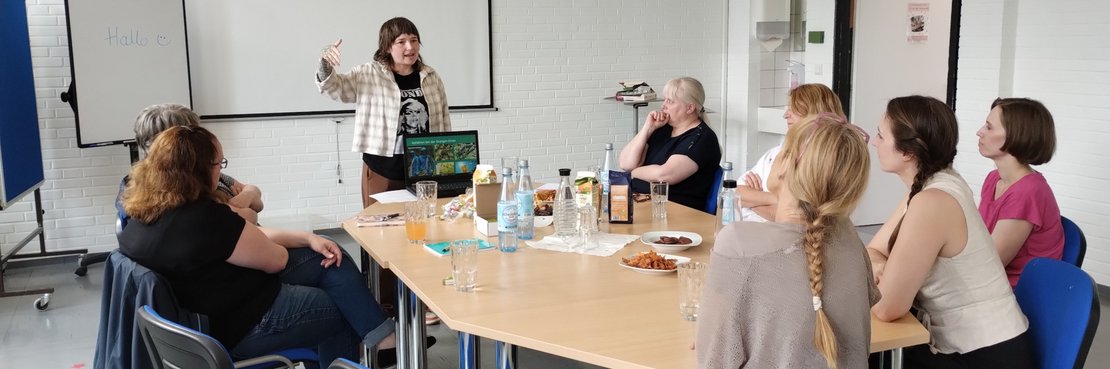 Eine Gruppe von Menschen sitzt an einem Tisch und hört einer Frau zu, die vor ihnen steht. Auf dem Tisch stehen Getränke und Snacks.